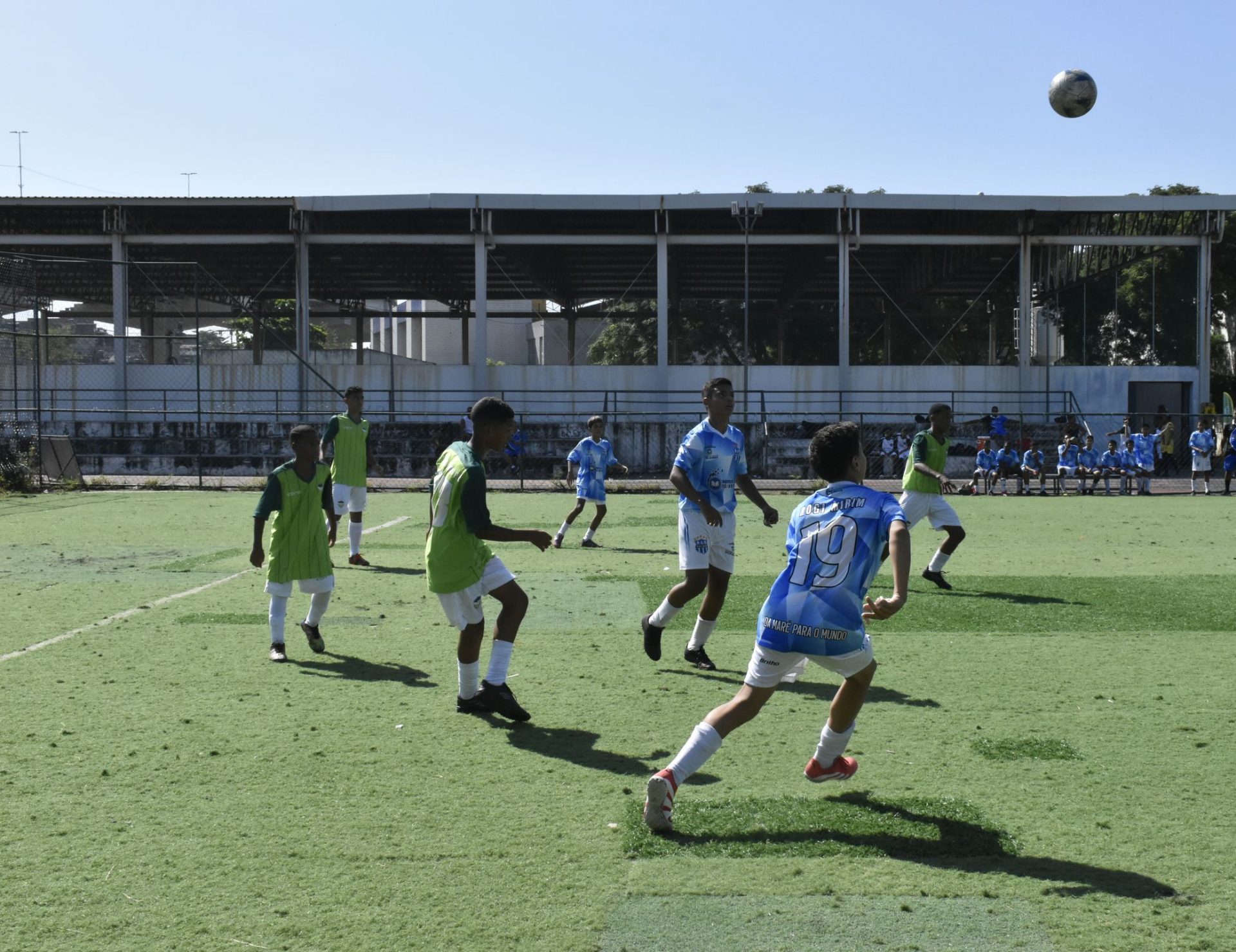 Garotos de colete verde e de camisa azul numerada disputam uma bola alta num campo de grama sintética com linhas marcadas; ao fundo, a arquibancada coberta e os reservas no banco.