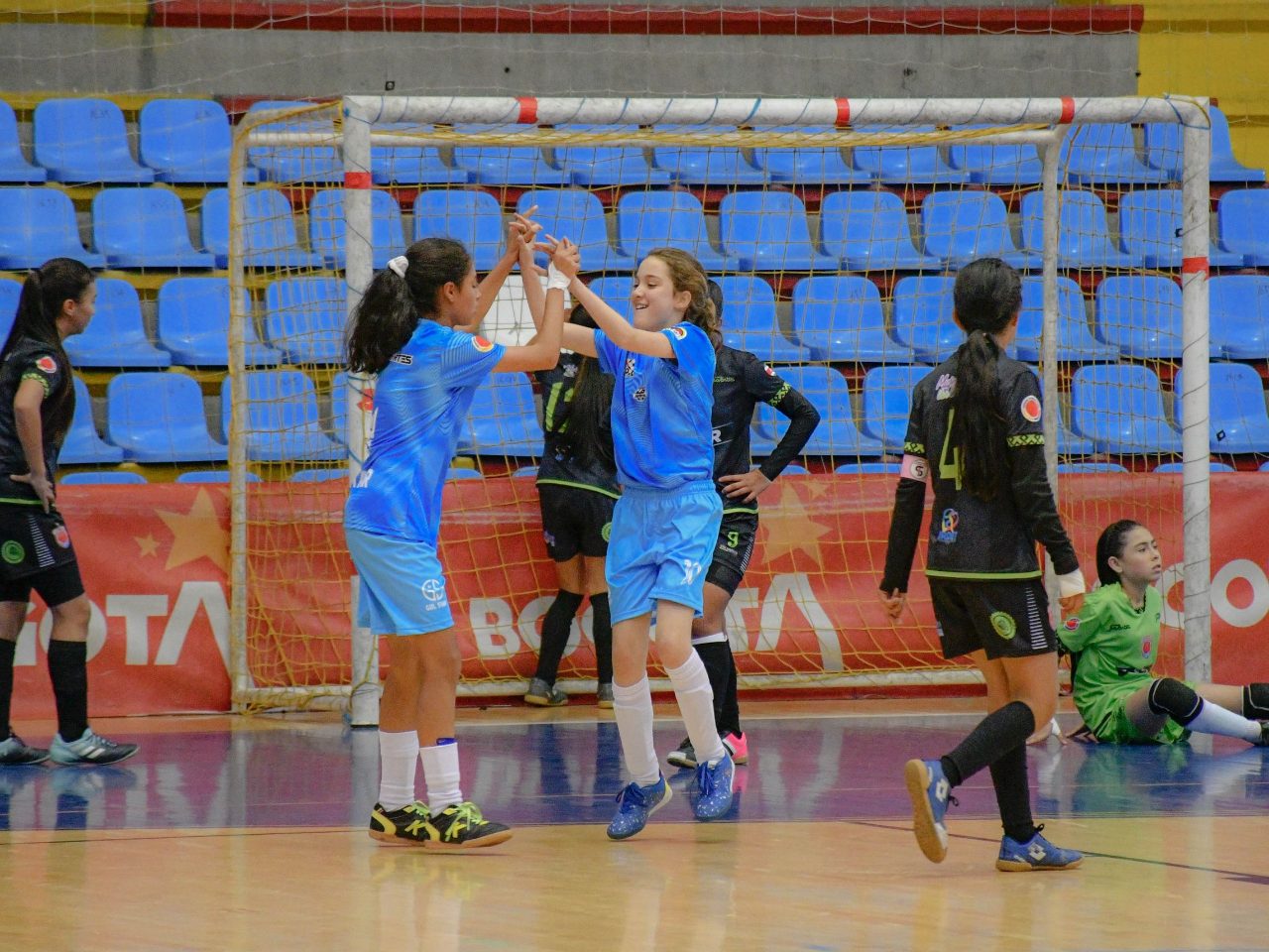 Meninas de um time de futsal batem as mãos comemorando um gol num ginásio, com a trave e as cadeiras azuis da arquibancada ao fundo.