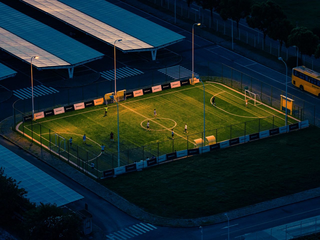 Vista do alto de um campo society de grama sintética iluminado ao anoitecer, com linhas marcadas, alambrado e jogadores em partida.